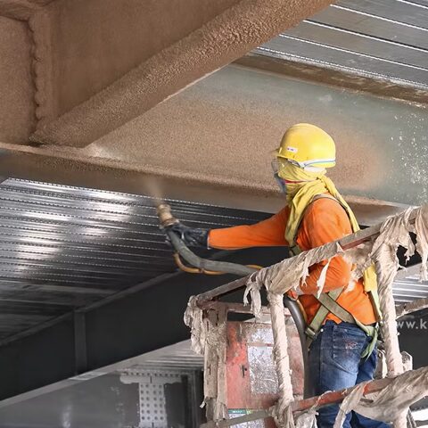 Worker applying fireproofing spray coating to a steel structure inside an industrial facility.